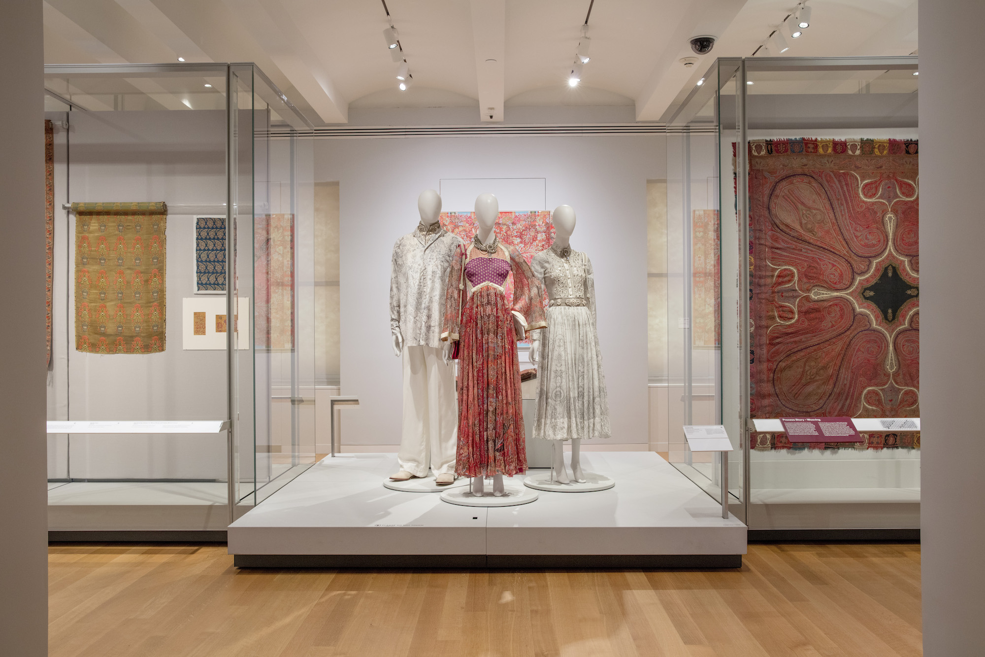 Three manikins stand in the middle of a gallery, wearing red and white clothing with paisley patterns. Displayed on both sides of the manikins are large textiles with paisley motifs.
