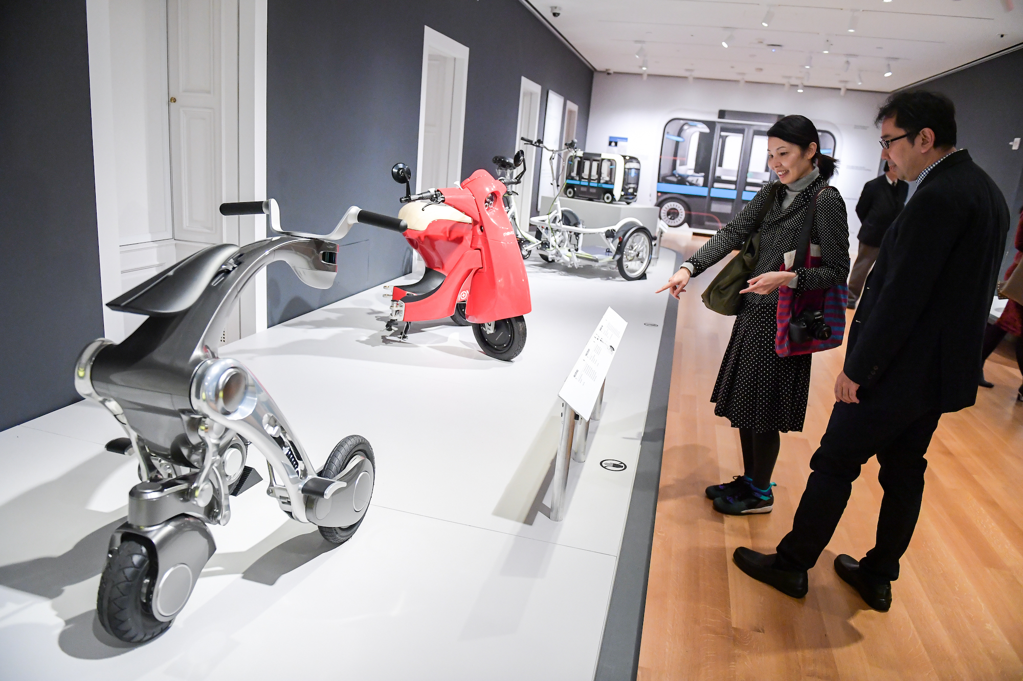 A woman in a gallery points to a futuristic, chrome-colored scooter, which is on display alongside a folded red scooter, a large bike, and a vehicle that looks like a small bus.