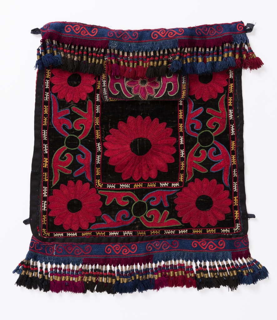 Image features a pair of bags with a flap opening in the center. Black velveteen fronts, embroidered in a large-scale floral design in red with outlines in blue and green; inner and outer borders with a geometric design in yellow and off-white. Woven bands in indigo blue at top and bottom, embroidered with a scrolling design in two shades of red, ending in fringes in dark red, blue and black, wrapped with gold metallic yarns. Backing is red cotton with a discharge print in white of two paisleys, a star and cresent moon. Hanging loops at upper corners. Please scroll down to read the blog post about these objects.