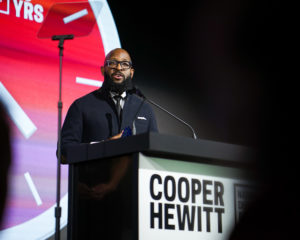 A man stands at a podium that says Cooper Hewitt, holding a blue trophy and appearing to give a speech. Behind him is a large clock with a pink and purple gradient.