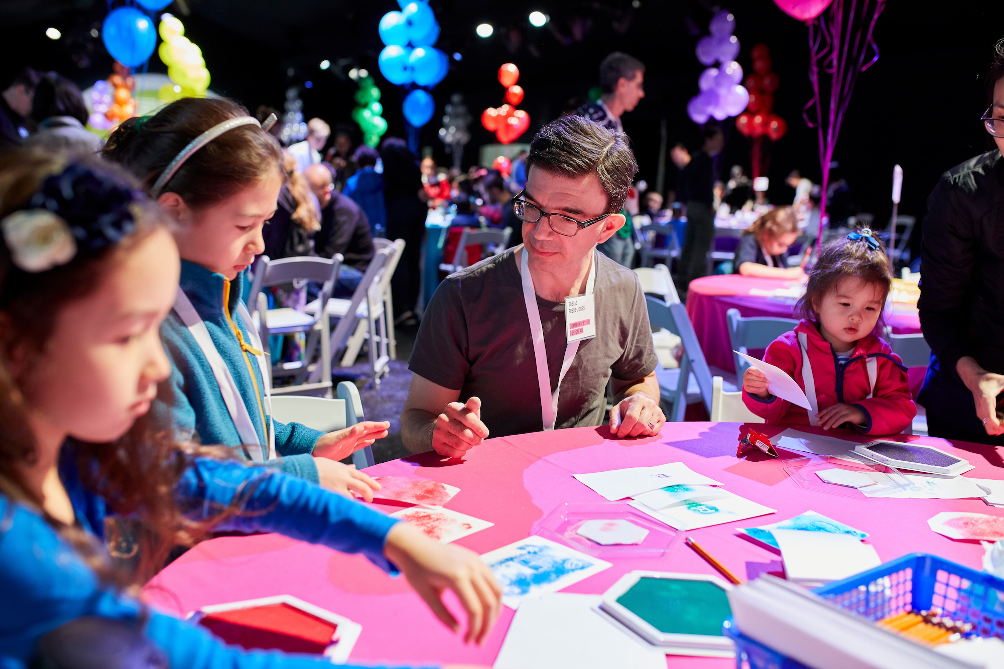 People gather at a table, in a festive room surrounded by balloons, creating postcards using bright colors.