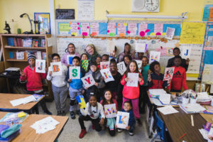 Kids gather in a classroom, each holding a piece of paper with colorful typography, smiling for a group shot.