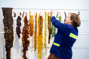 Julia, a fair-skinned woman with a blonde bob, smiles as she hangs strips of colorful seaweed from a clothesline. She wears a blue worker's jacket that says Department of Seaweed on the back.
