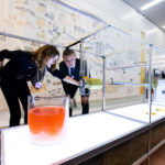 A designer and a visitor lean in to look at an experimental lab machine, where a glowy vat of orange goop sits near a roller or printing machine with holes cut out of it