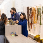A woman explains the concept of the Department of Seaweed to a visitor. Behind her hang strips of dried seaweed that is green, gold, and brown. The woman is standing at a table where a small seaweed structure rests.
