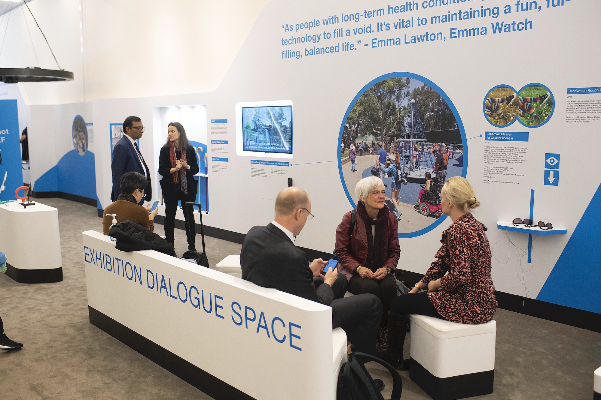 A group of people are gathered in an area labeled “Exhibition Dialogue Space” next to a blue and white wall full of images, video, and text.