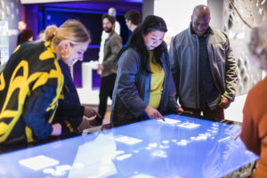 A group of people gather around a large horizontal digital screen, using their fingers to interact with it.