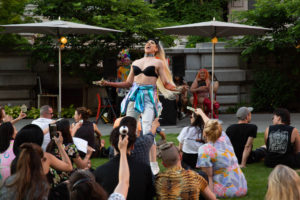 A drag queen with long hair, a black bikini top, elbow length gloves, and an iridescent jacket tied around her waist performs on a grass lawn. A crowd gathered around her are taking photos.