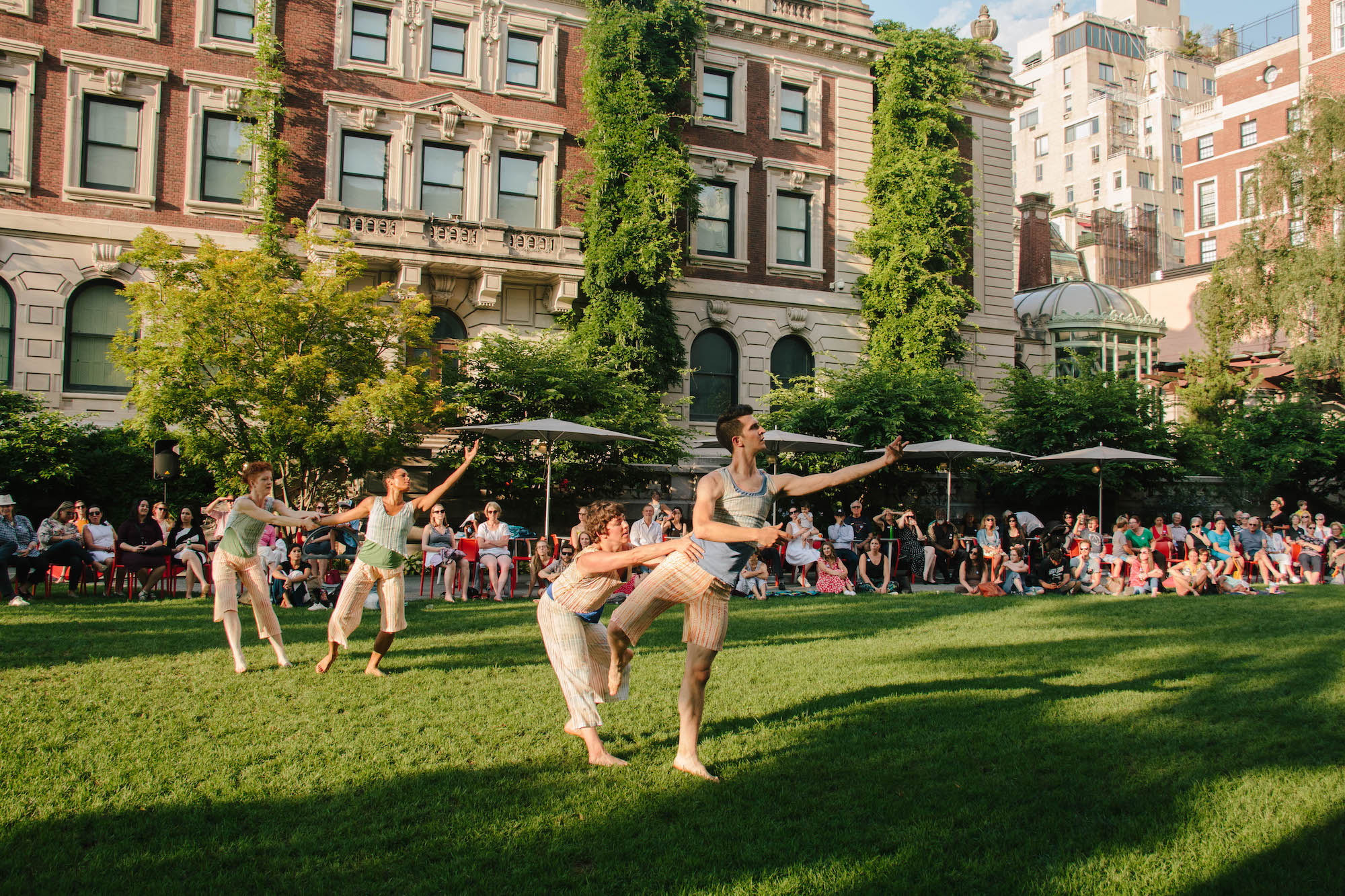 Two pairs of dancers perform on a grass lawn in front of a large mansion. An audience watches from behind, sitting on orange chairs.