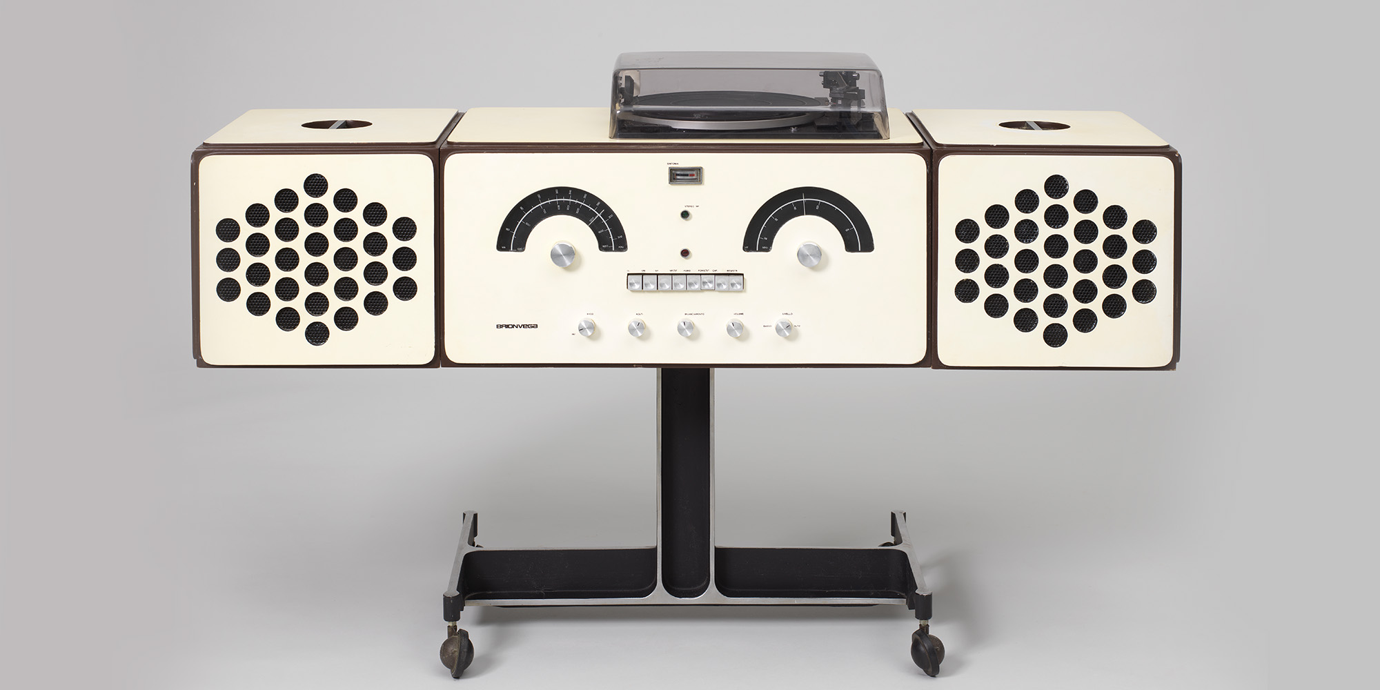 Mid-century stereo console. The unit is made of three sections. The center rectangular section houses radio controls arranged to resemble a human face and a record player. It is flanked by detachable box shaped speakers.