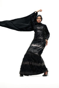 Raşit Bağzıbağlı, a glamorous model with a rich skin tone, poses with one arm above her head, her knees angled in an awkward-but-chic way that screams "high fashion." She wears an all black ensemble, including a head wrap, a long sequined dress, and tall black high heels. Extending from her lifted arm, caught in the wind like a flag, is a black silky shawl.