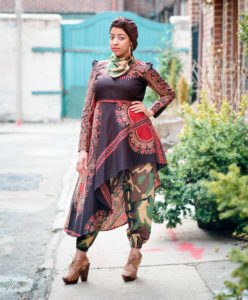 A cool influencer with a medium skin tone shows off her street style. She has one hand on her hip and stares unsmiling at the camera. She wears a brown head wrap, gold hoop earrings that are so large they brush her shoulders, a camo-print scarf around her neck, an eggplant-color dress with an asymmetrical hem with long sleeves and red and gold detailing, camo-print pants, and taupe open toe lace-up platform high heel shoes.