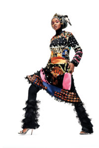 Dian Pelangi, a beautiful model with a rich skin tone and angular features, models a tunic layered over leggings. Her head is wrapped in a black, green, and white cloth with small white circles outlining her face. The tunic is black and decorated with many complicated appliques and beads in hues, including pink ribbons that encircle her wrists. The tunic ends just above her knees. The leggings she wears are black and decorated with layers of black fringe or tassels. The ensemble concludes with glamorous open-toe silver stilettos.