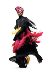 Dian Pelangi, a model with a medium rich skin tone and very high sharp cheekbones, faces the camera and lifts one leg, setting her tassel-covered ensemble into motion. She wears a pink and black headwrap. Her garment, which covers her from head to toe, is extremely avant-garde. It is composed of alternating layers of a fine black mesh, long yellow tassels, long black tassels, and long pink tassels. She wears experimental-looking teal eye shadow and her eyes are narrowed.