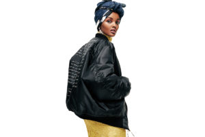 Céline Semaan Vernon, a beautiful and elegant model with a rich skin tone, twists her head backward to look at the viewer. She wears a blue headwrap, a long, tight yellow dress with floral lace, dangly diamond earrrings, and a black bomber jacket. On the back of the jacket is Arabic text written in white. She is wearing red eyeshadow and her mouth is slightly parted.