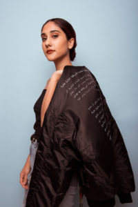 A young woman with a rich olive skin tone tosses over her shoulder a black flighter jacket with white Arabic text on the back. Her hair is tied back in a bun and has a lone red highlight. She has dramatic eyebrows, cat eye eyeliner, and is wearing lipstick. A trio of stud-shaped earrings sparkle on her ear lobes.