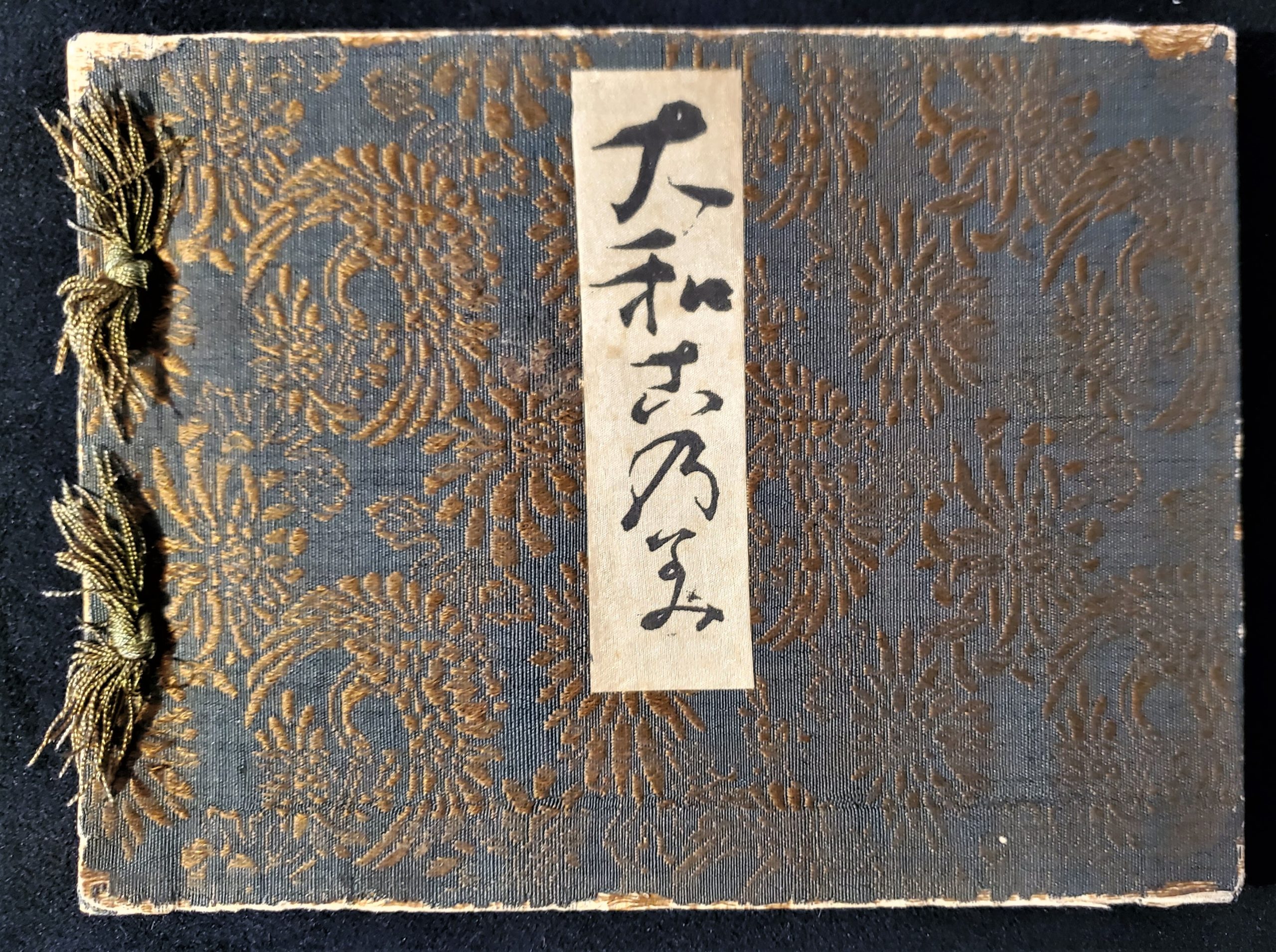 Image features the cover of the 1905 Yamanaka & Co. furnishings trade catalogue covered in green and gold silk brocade and bound on the left with gold silk threads. A vertical rectangular white paper panel with Japanese characters is in the center of the cover. Please scroll down to read the blog post about this object.