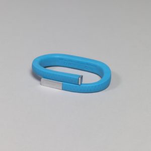 Image features a slim blue band, rectangular in section, coiled to fit around the wrist. There is a zig-zag pattern on the rubber surface, a thin metal cap at one end, and a metal strip with the word "JAWBONE" at the other. Please scroll down to read the blog post about this object.