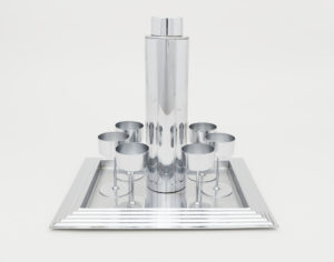 Image features a polished chrome-plated cocktail service consisting of tall cylindrical cocktail shaker and lid, a rectangular tray with stepped rim; and six cylindrical cups with slender stems and circular feet. Please scroll down to read the blog post about this object.