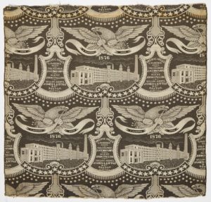 Image features a woven textile that serves as a commemorative of the US centennial while promoting the textile manufacturer Pacific Mills of Lawrence, Massachusetts. Woven in black and cream, the composition has a bold graphic quality and depicts a broadly soaring eagle over a large modern factory enclosed in curvilinear frame. In between each factory is a plaque identifying the Treasurer, Agent and Selling Agent of the business. Please scroll down to read the blog post about this object.