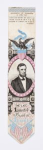 Image features a bookmark or stevengraph with a portrait medallion of Abraham Lincoln surmounted by an eagle perched on a shield flag that holds a banner in its beak that reads "E Pluribus Unum." Inscription at top reads: "Assassinated at Washington 14 April 1865," and just below another inscription: "I have said nothing but what I am willing to live by. And if it be the pleasure of Almighty God. To die by. (A Lincoln). Please scroll down to read the blog post about this object.