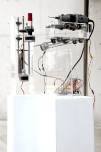 Machine on white plinth made of metal, clear plastic, and wires. The machine holds up a red lipstick