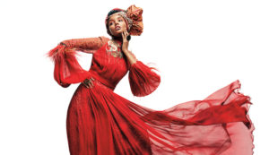 A glamorous model wearing a flowing red gown and a large hairwrap strikes a pose