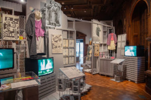 View of gallery with industrial-inspired installation featuring clothing and videos