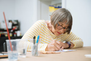 A woman with grey hair and a yellow striped shirt colors on a table.
