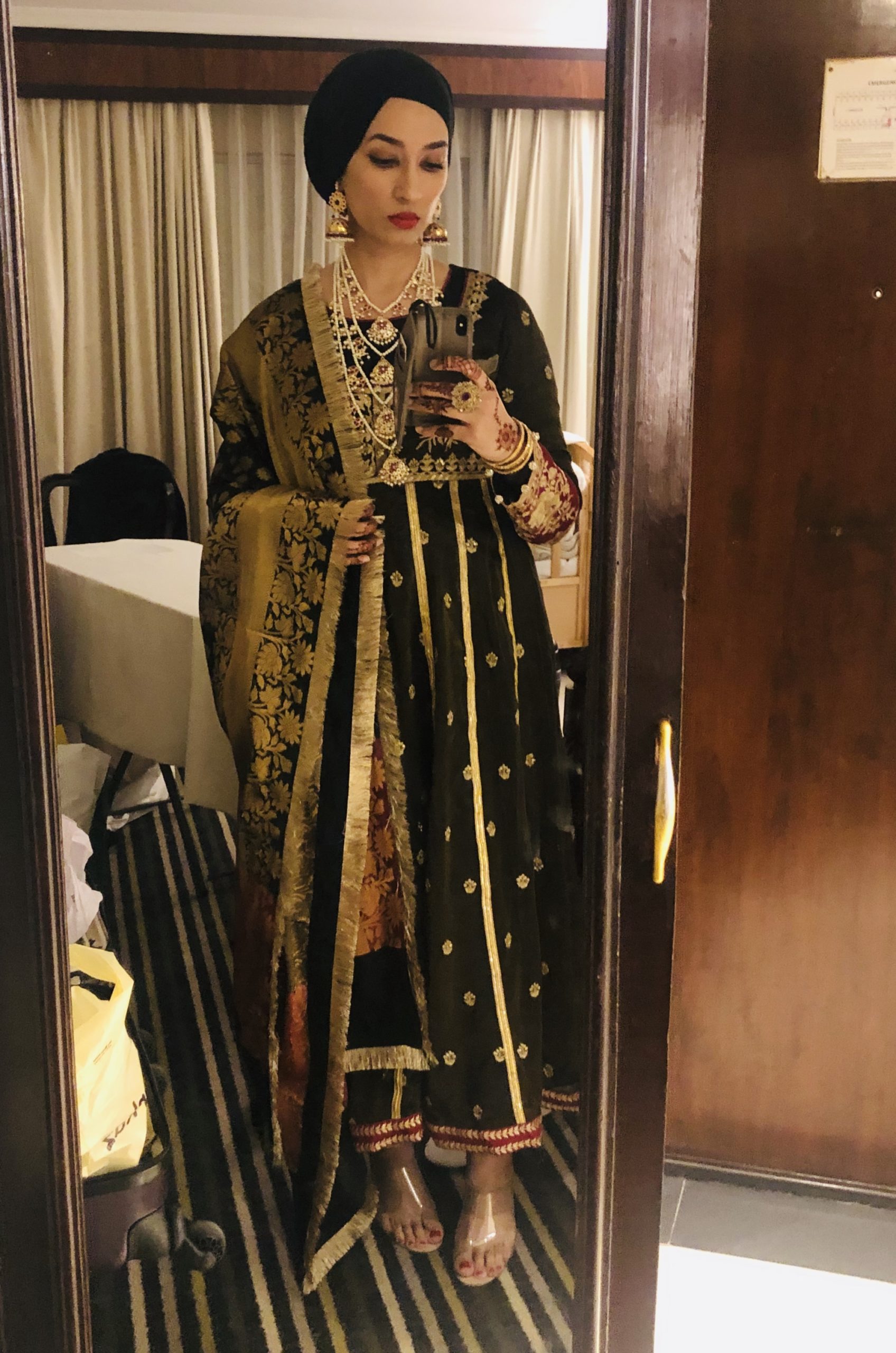 Mirror selfie. Saba Ali gazes at self through phone camera, dressed in extremely elegant formal wear. She wears a long black abaya with intricate gold embroidery and a black headwrap. She wears many gold necklances, large gold earrings, shoes made from clear plastic, and her hands are decorated with Henna. She appears to be in a hotel room.