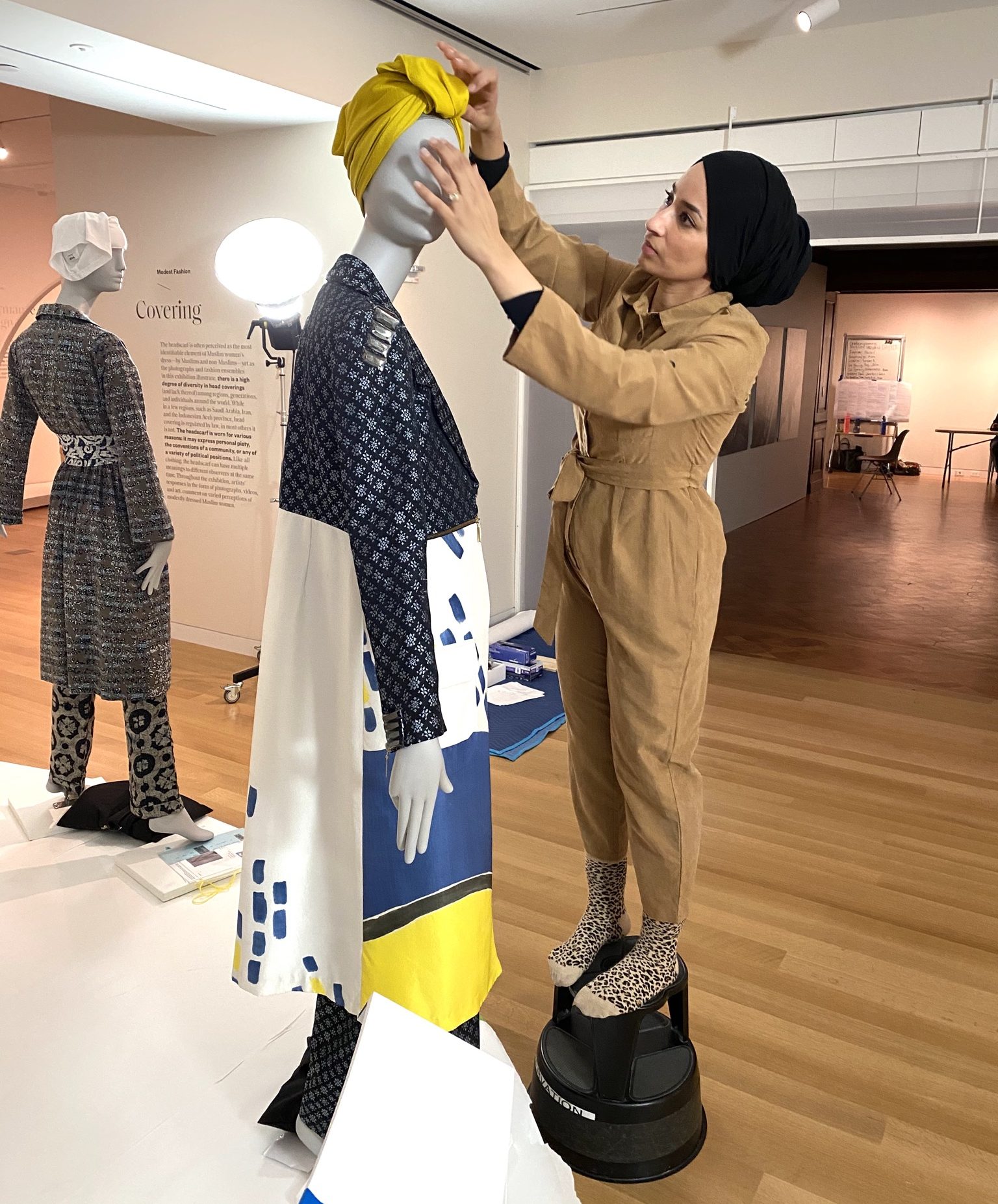 Behind the scenes at Cooper Hewitt. Saba Ali, a hip woman wearing a taupe jumpsuit and a black head wrap, stands on a stool so as to adjust and style a yellow headdress on a mannequin wearing a high-fashion ensemble