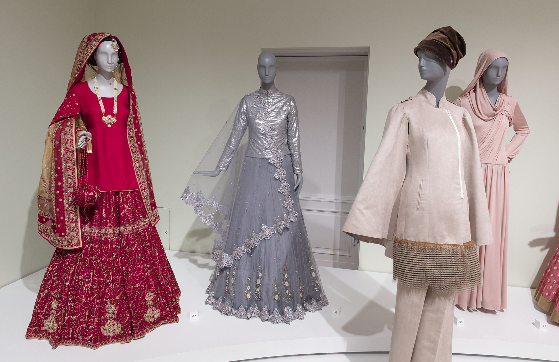 Saba's dazzling red wedding dress with ornate golden embroidery and a long matching shawl. The mannequin is also styled with a delicate golden chain leading from its ear to the nostril (a nosering) and a red handbag with a long beaded chain attached to a spherical pouch. To the right of Saba's wedding ensemble are three more looks: a silver gown, a pale pink tunic and matching pants with a fringe made from taupe feathers, and a draped pink jersey number with a built-in headwrap.