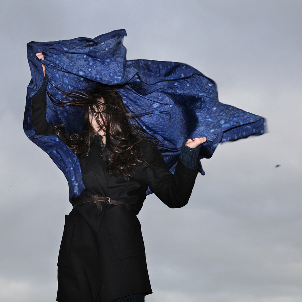 A model whose long dark hair obscures her face lifts above her head a navy blue scarf speckled with an irregular pattern created by raindrops. The sky behind her is gray and stormy.