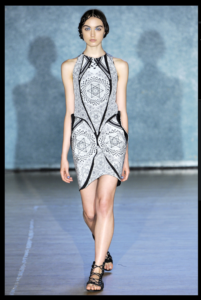 A model walks down a runway wearing a short sleeveless dress with a white and black pattern featuring tessellating six pointed stars within hexagons. She has short brown hair tied into braided crowns and wears black gladiator sandals.
