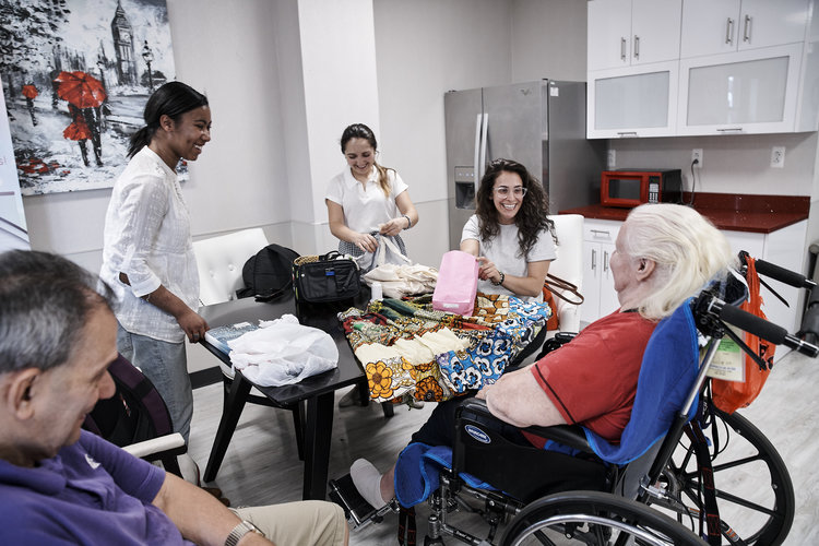 Three smiling young women in white shirts stand at a table with an outstretched fabric sample with a colorful print. Across from them sits a woman with white hair who is seated in a wheelchair. Slightly out of frame is a smiling gray haired man looking on at the proceedings. Behind the women is a fridge and a counter space suggesting they are in the shared cafe area of an office or school building.