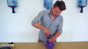A man creates a purple bioplastic vase. Three water jugs hang on the wall behind him.
