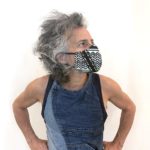 Adi, a man with wild gray hair, poses with his hands at his hips. He wears a blue tank top and a fabric mask made from fabric with an ornate black and white design