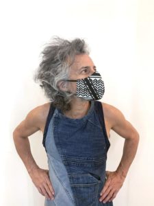 Adi, a man with wild gray hair, poses with his hands at his hips. He wears a blue tank top and a fabric mask made from fabric with an ornate black and white design