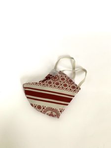 View of a cloth mask folded and placed on its side. The mask is made from fabric with a red and white pattern. The pattern features six-pointed stars within hexagons. The pattern is interrupted by bold red stripes. The mask has elastic bands to be fixed behind the wearers ears