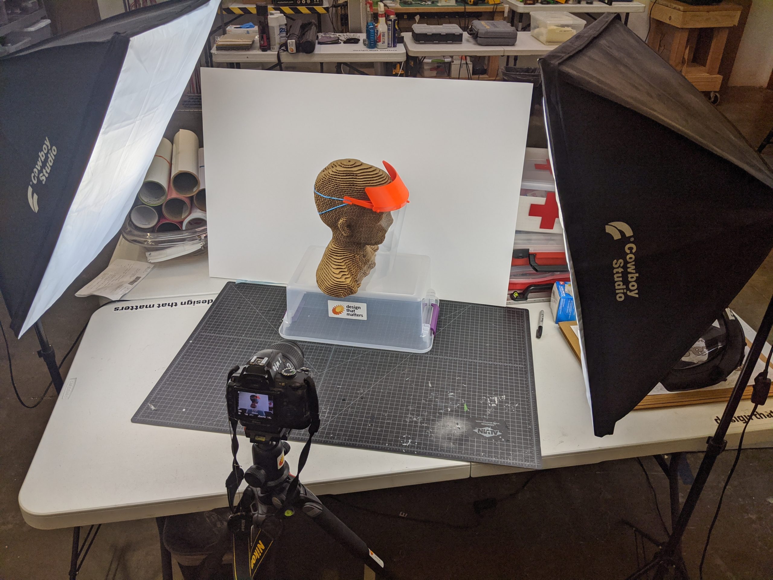 3D printed form of a mannequin head, wearing a face shield and propped up on a clear box, is photographed in a studio. The face shield features a bright orange headband to which is attached a large piece of clear plastic