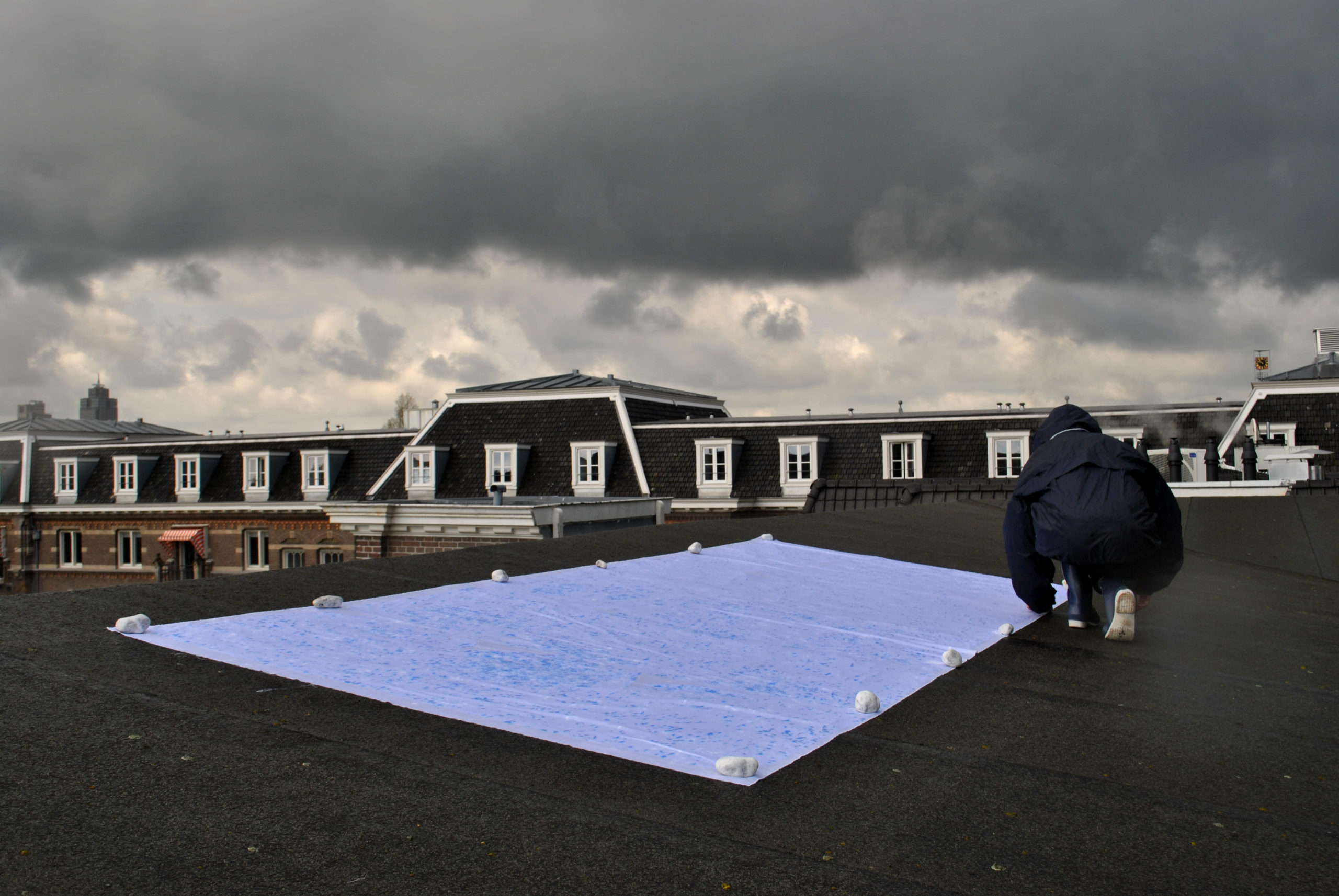 On a stormy day, a figure in rain gear lays out a sheet of plastic on a field on the roof of a building. The plastic is anchored with white stones, and the facade of a grand house can be seen in the near distance.