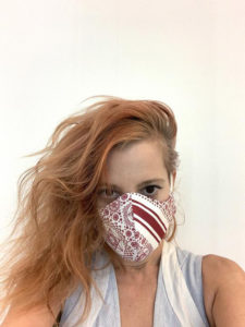 Idi Gil, a young woman with vibrant orange hair, takes a selfie while wearing a facemask. The facemask is made from a white cloth with a red pattern.