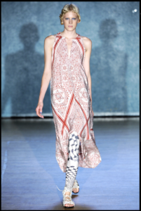 A model with blonde hair walks down the runway in a long, flowing dress with a red-and-white pattern featuring tessellating six-pointed stars. She walso wears leggings with an abstract black and white pattern and white gladiator sandals.