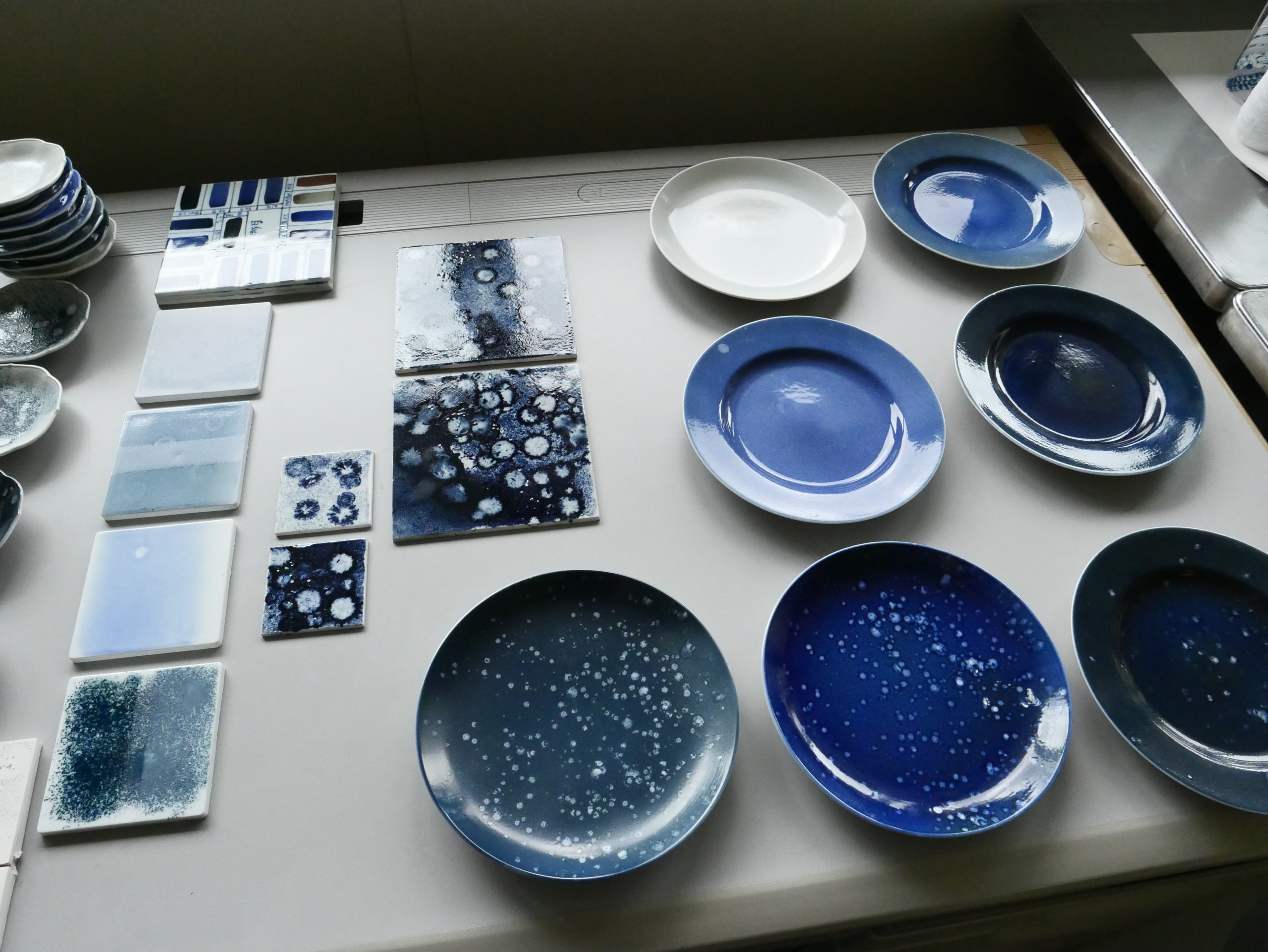 An array of porcelain plates and sample material squares in blue tones ranging from deep navy to light blue to blue-green. These materials are faintly speckled with white dots in an irregular pattern, evoking the pattern formed by fallen raindrops.