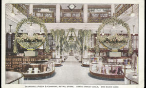 This postcard depicts a swanky department store in another era. Within a palatial interior, retail displays are ornamented with two identical giant wreaths. On the upper level of the building, a hanging clock marks the hour between wooden display cases of ceramics. The photo is captioned Marshall Field & Company, Retail Store, State Street Aisle, One Block Long.