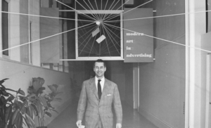 Black and white photograph of man in a suit, standing in a hallway, with a sign behind him that says modern art in advertising