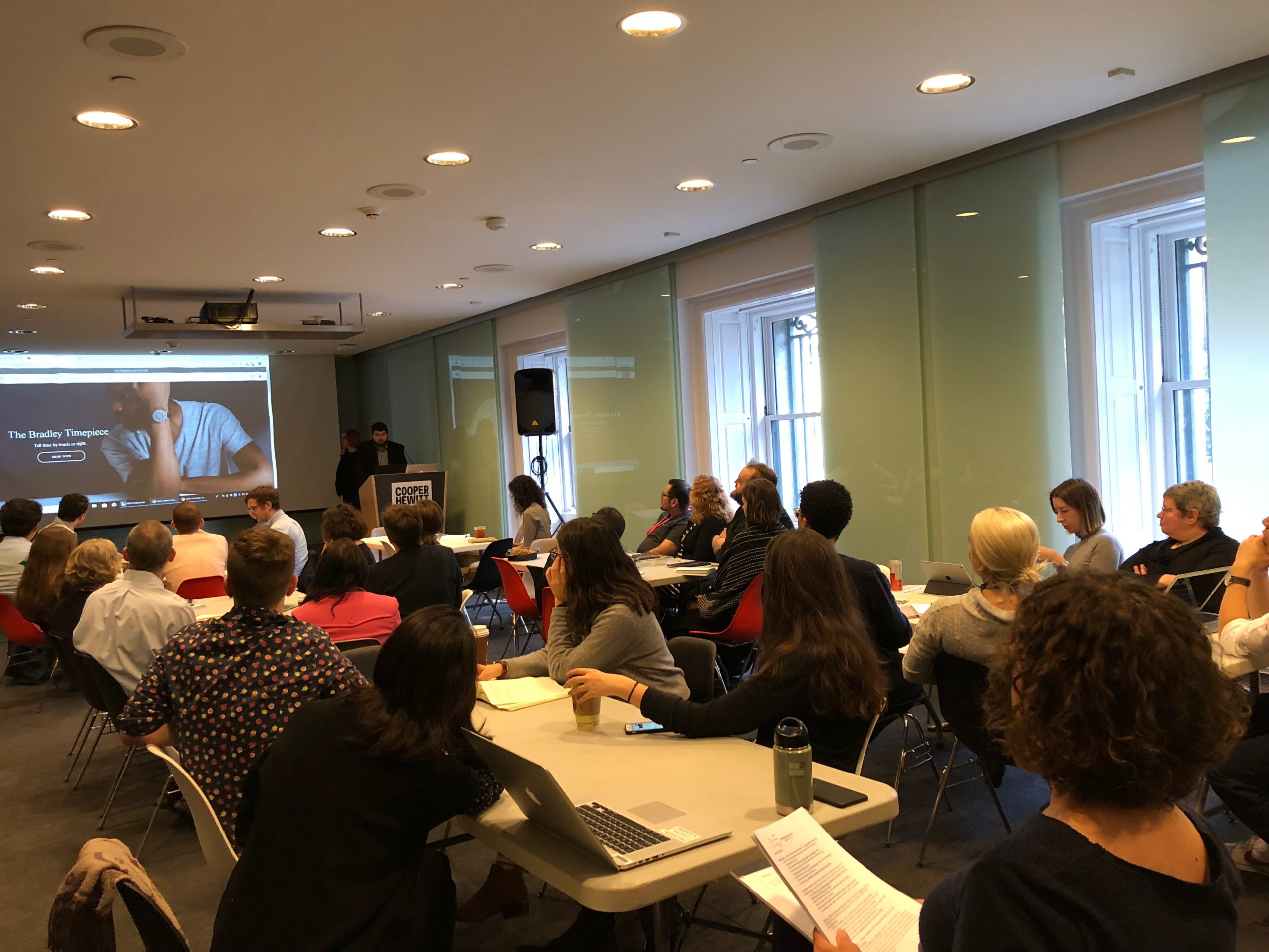 Many people, CH staff, seated at tables facing a projector screen which displays an image of the Bradley timepiece. Anna and Sina stand on stage at the front of the room.