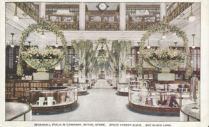 This postcard depicts a swanky department store in another era. Within a palatial interior, retail displays are ornamented with two identical giant wreaths. On the upper level of the building, a hanging clock marks the hour between wooden display cases of ceramics. The photo is captioned Marshall Field & Company, Retail Store, State Street Aisle, One Block Long.
