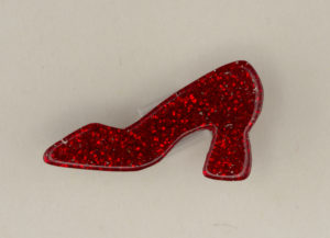 A smooth plastic pin in the form of a high-heeled shoe in profile with the toe pointing left. Shoe is a block-heeled, classic vintage style pump that is translucent, sparkly, ruby red with embedded red glitter.