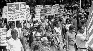 In this historic black and white photo, a crowd of African American civil rights protesters is gathered. Many hold signs that say "We march for higher minimum wages coverage for al workers now!" Another man carries an American flag.